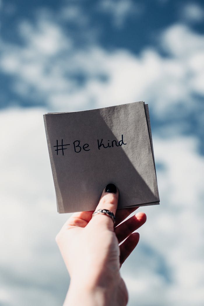 A hand holding a note with '#Be kind' inscribed, set against a bright sky.